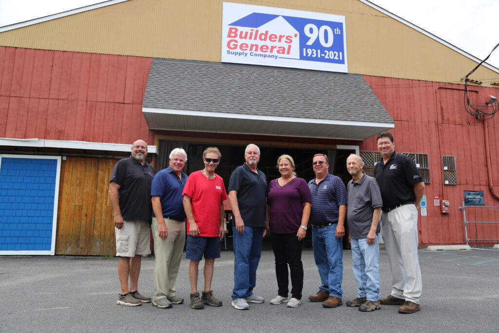 Builders’ General employees standing in front of New Jersey building supply store. Sign above saying "Builders’ General supply store 90th" for their 90th anniversary.