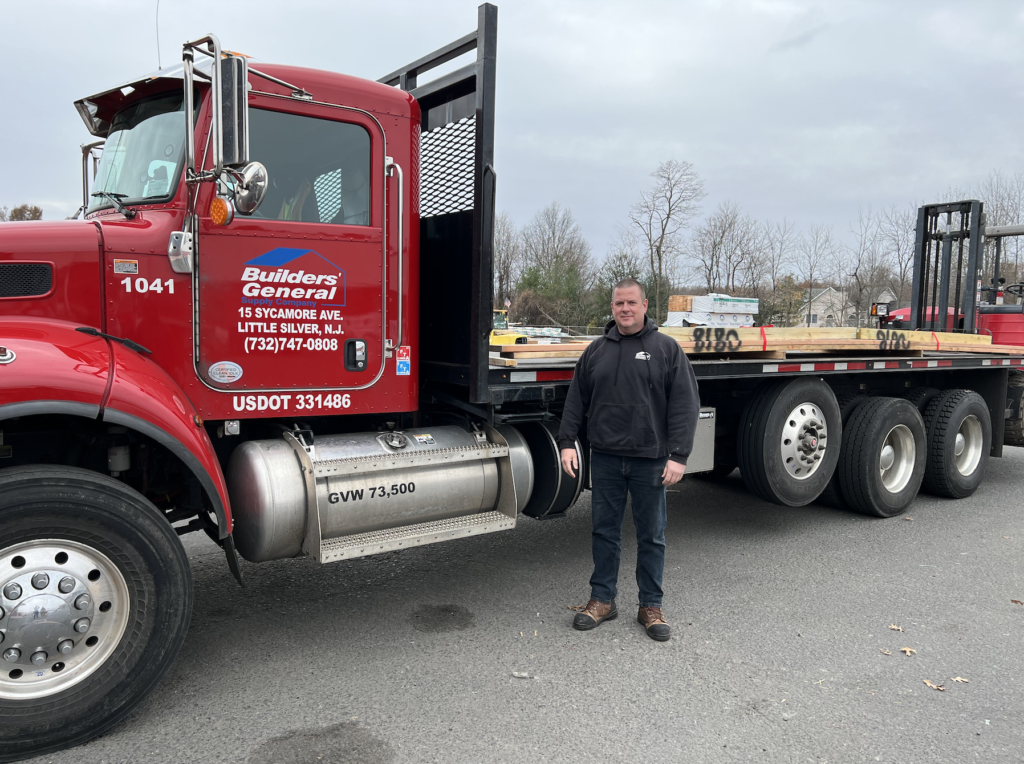 Tim Bradley stands in front of a red Builders’ General truck. He is wearing a black sweatshirt, dark blue jeans and brown work boots. There is lumber on the back of the truck.