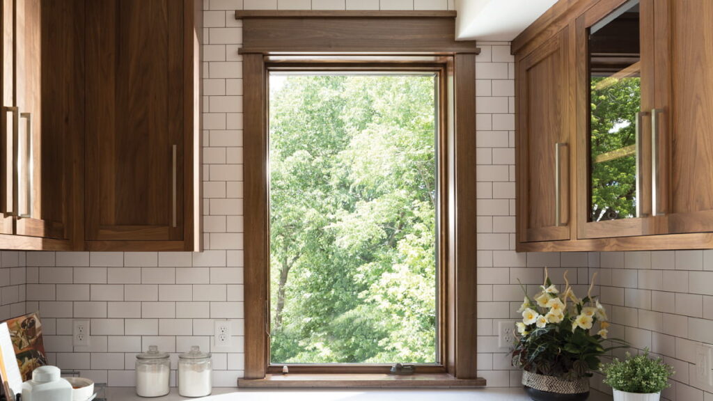 A 400 series Andersen window in brown is centered in a kitchen looking outside to lush greenery. There are brown kitchen cabinets on both sides on a white tiled wall with glass containers of sugar and flour and a few plants.