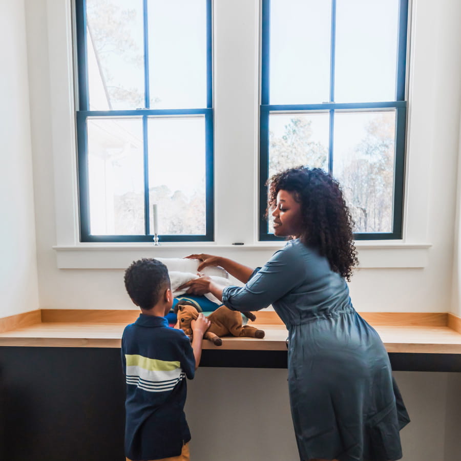 A woman wearing a blue dress and her young son are standing near a wood-top counter in front of two large, black Andersen 400 Series windows.