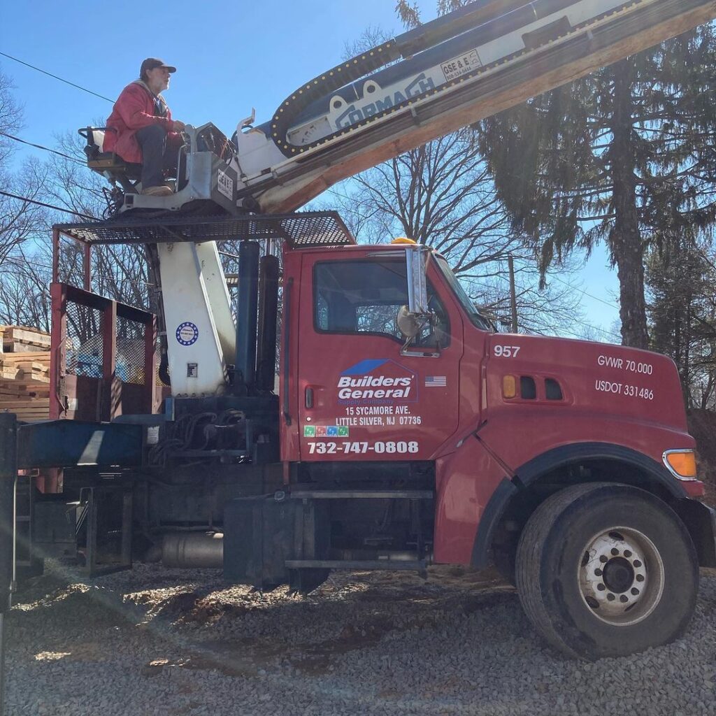 A boom truck is placing lumber on a jobsite. The person on the boom truck is wearing a red jacket and black pants. The truck is red with the Builders’ General logo on the side.
