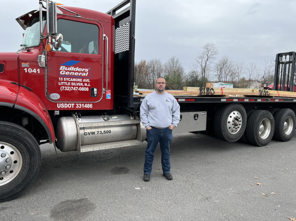 Employee John Powers standing next to a Builders' General delivery truck loaded with building products. He is wearing a grey crewneck, dark blue jeans and grey work boots. He has both hands in his pockets. 